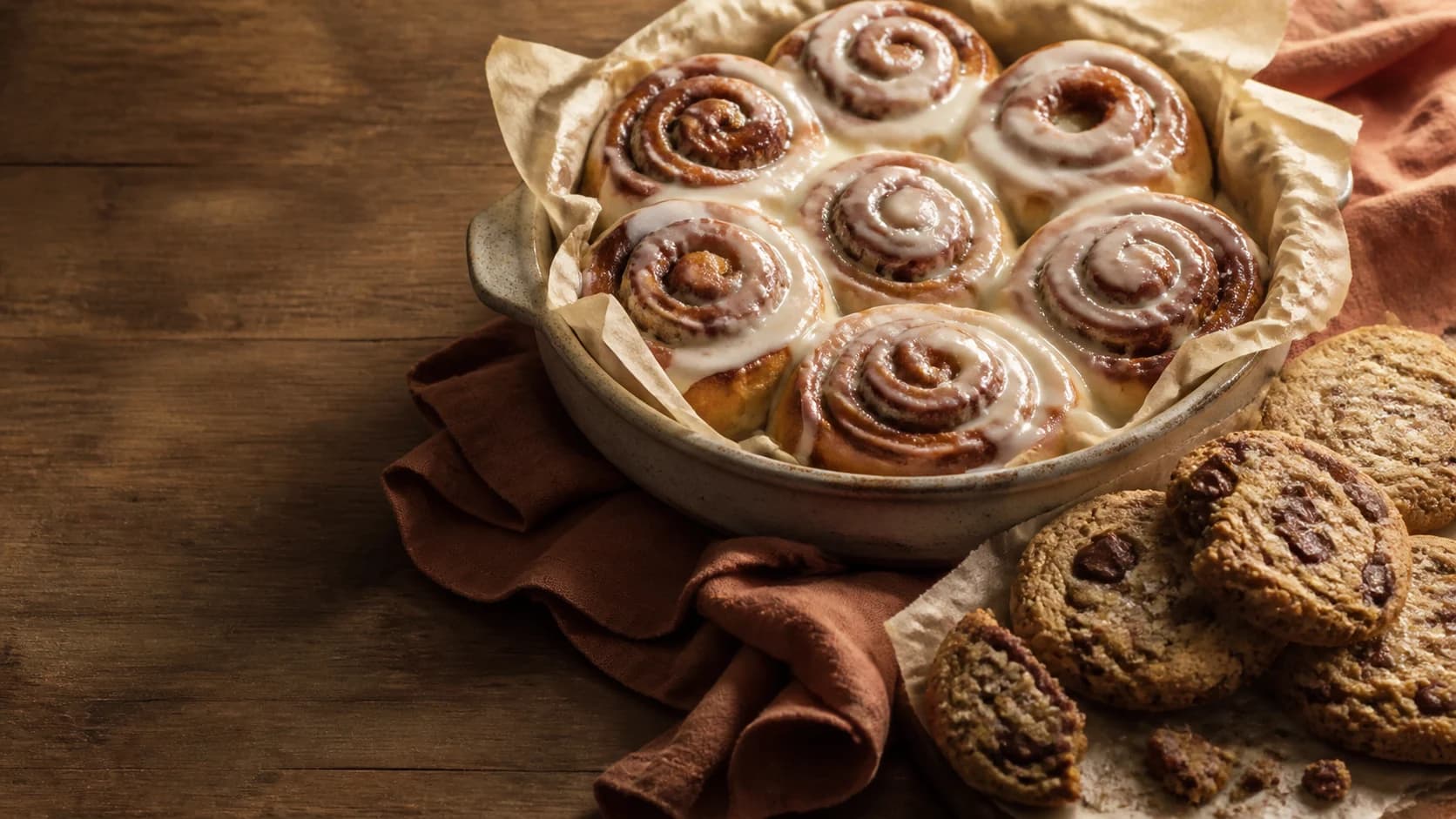Glazed cinnamon rolls and chocolate chunk cookies arranged on a warm wooden table.